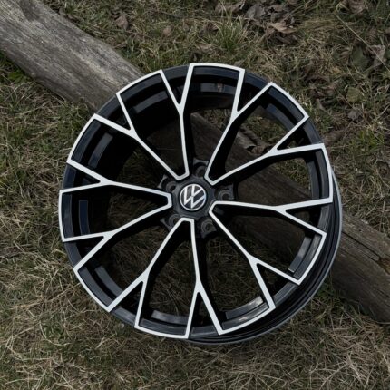 Black alloy car wheel with a white multi-spoke design and VW center cap, resting on grass beside a wooden log.