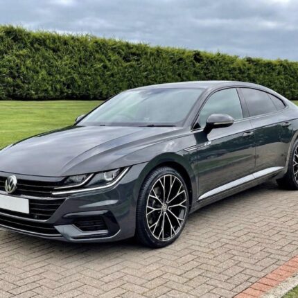 Front-three-quarter view of a dark gray Volkswagen sedan parked on a brick driveway with a green hedge in the background.