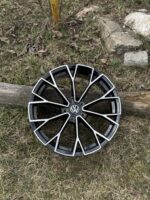 Black and silver VW alloy wheel resting on grass beside a wooden rail and stones.