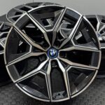 Close-up of a glossy black BMW alloy wheel with angular spokes and chrome trim, featuring the blue-and-white BMW emblem at the center on a textured surface.