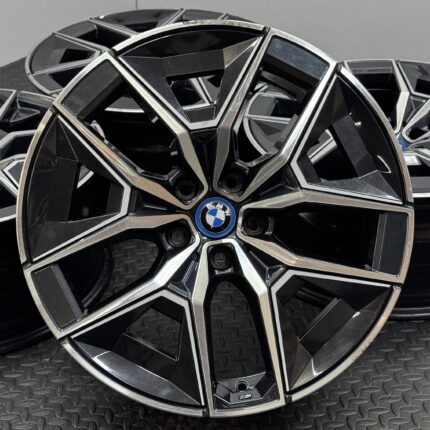 Close-up of a glossy black BMW alloy wheel with angular spokes and chrome trim, featuring the blue-and-white BMW emblem at the center on a textured surface.
