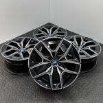 Stacked BMW alloy wheels with black and silver multi-spoke design, centered blue BMW badges, on a textured metal floor.