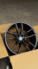 Black BMW alloy wheel with a multi-spoke design placed inside an open cardboard box on a rough pavement surface.