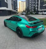 Teal BMW sports car with a black roof parked in a modern residential courtyard between tall buildings, reflecting the surrounding facades in its surface.