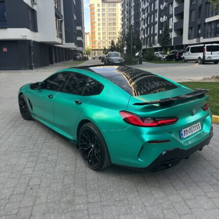 Teal BMW sports car with a black roof parked in a modern residential courtyard between tall buildings, reflecting the surrounding facades in its surface.