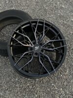 Black multi-spoke alloy wheel lying on a gravel surface with a tire in the background, showcasing an intricate spoke design.