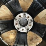 Close-up of a vehicle wheel hub with six lug holes, set in a circular metal center, surrounded by a black plastic wheel cover on an OSB board background.