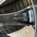 Close-up of a black car wheel/rim with a tire, showing engraved numbers and a small emblem on the rim edge.