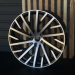 Close-up of a glossy black and silver alloy wheel with the Audi center cap, against a wooden slat backdrop missioned as a product shot.