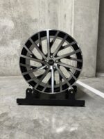Silver and black multi-spoke alloy wheel mounted on a display stand in a gray concrete space.