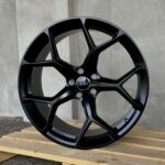 Close-up of a glossy black alloy car wheel with five spokes, resting on a wooden pallet; Audi center cap visible.