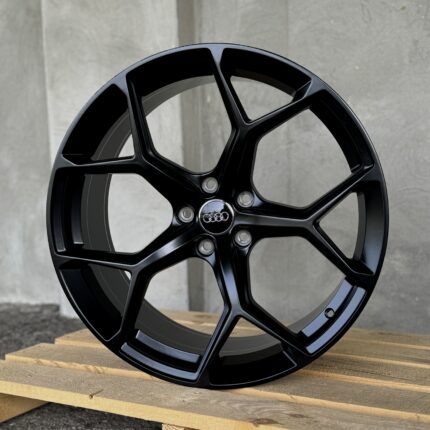 Close-up of a glossy black alloy car wheel with five spokes, resting on a wooden pallet; Audi center cap visible.