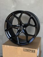 Black glossy alloy wheel rim with the Audi emblem, mounted on a cardboard box against a gray wall.