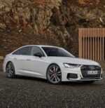 White Audi sedan parked on gravel with a rocky hillside and a vertical wooden slat barrier in the background.