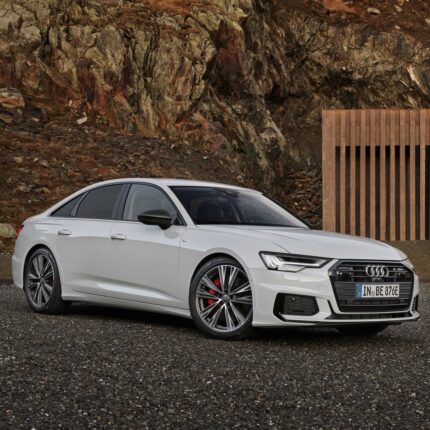 White Audi sedan parked on gravel with a rocky hillside and a vertical wooden slat barrier in the background.