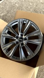 Audi alloy wheel with a multi-spoke design in a cardboard box on a gravel surface