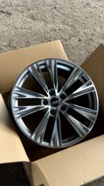 Silver multi‑spoke Audi alloy wheel in a cardboard box on dirt ground, center cap visible with Audi logo.