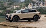 Beige Audi SUV parked on a rocky riverside with a hillside and cliff behind it.