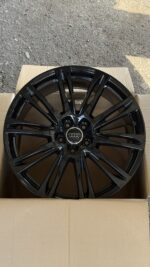 Black glossy multi-spoke Audi alloy wheel with central Audi emblem, placed inside a cardboard box on a gravel surface