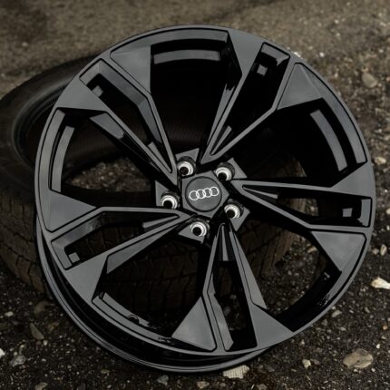 Close-up of a glossy black alloy wheel with the Audi logo at the center, resting on a gravel surface.