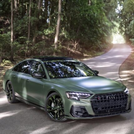 Front three-quarter view of a green Audi SUV parked on a winding forest road with trees on both sides.