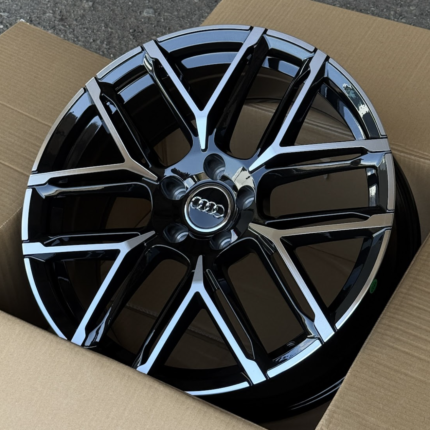 Glossy black Audi alloy wheel with silver accents, shown in a cardboard shipping box, front view