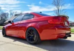 Red sports car parked on a suburban driveway, rear three-quarters angle, black roof and dark aftermarket wheels.