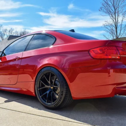 Red sports car parked on a suburban driveway, rear three-quarters angle, black roof and dark aftermarket wheels.