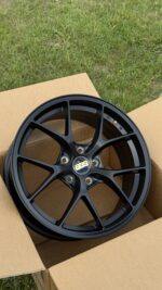 Black BBS alloy car wheel with a multi-spoke design, placed in a cardboard box outdoors on grass.
