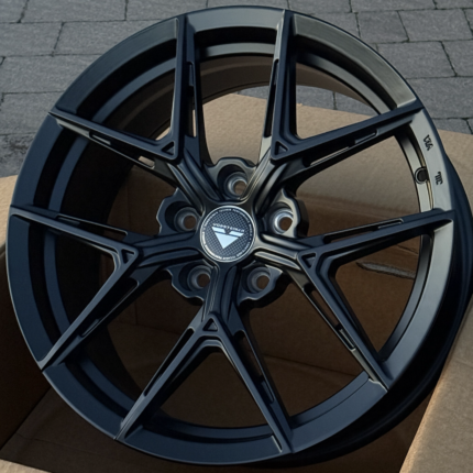Close-up of a glossy black Vorsteiner alloy wheel with a multi-spoke design and centered logo cap on a wooden surface.