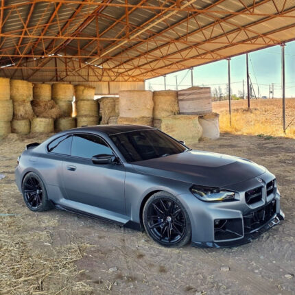 Matte gray BMW sedan with black rims parked next to stacked hay bales under a metal roofed barn in a rural setting.
