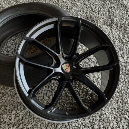 Close-up of a black alloy car wheel with a Porsche emblem, resting on a gravel surface outside a tire.