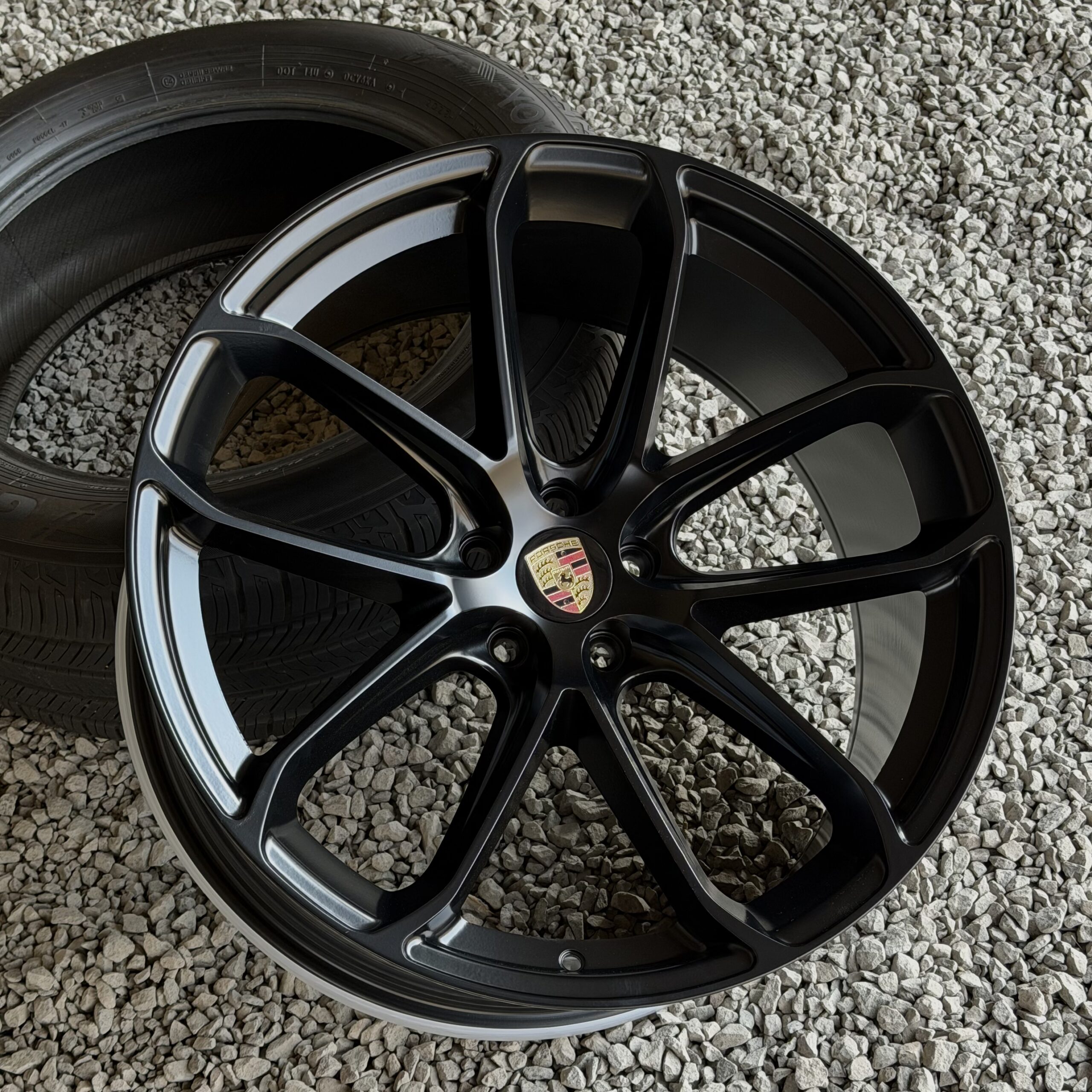 racingline_rfx-030_blackmachinedface Close-up of a black alloy car wheel with a Porsche emblem, resting on a gravel surface outside a tire.