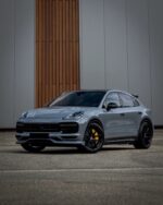 Silver Porsche SUV in a front three-quarter view with black wheels and yellow brake calipers, parked on a paved surface in front of a modern building with wood panel accents.