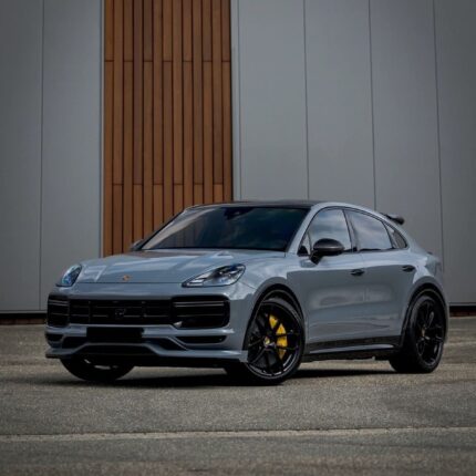 Silver Porsche SUV in a front three-quarter view with black wheels and yellow brake calipers, parked on a paved surface in front of a modern building with wood panel accents.
