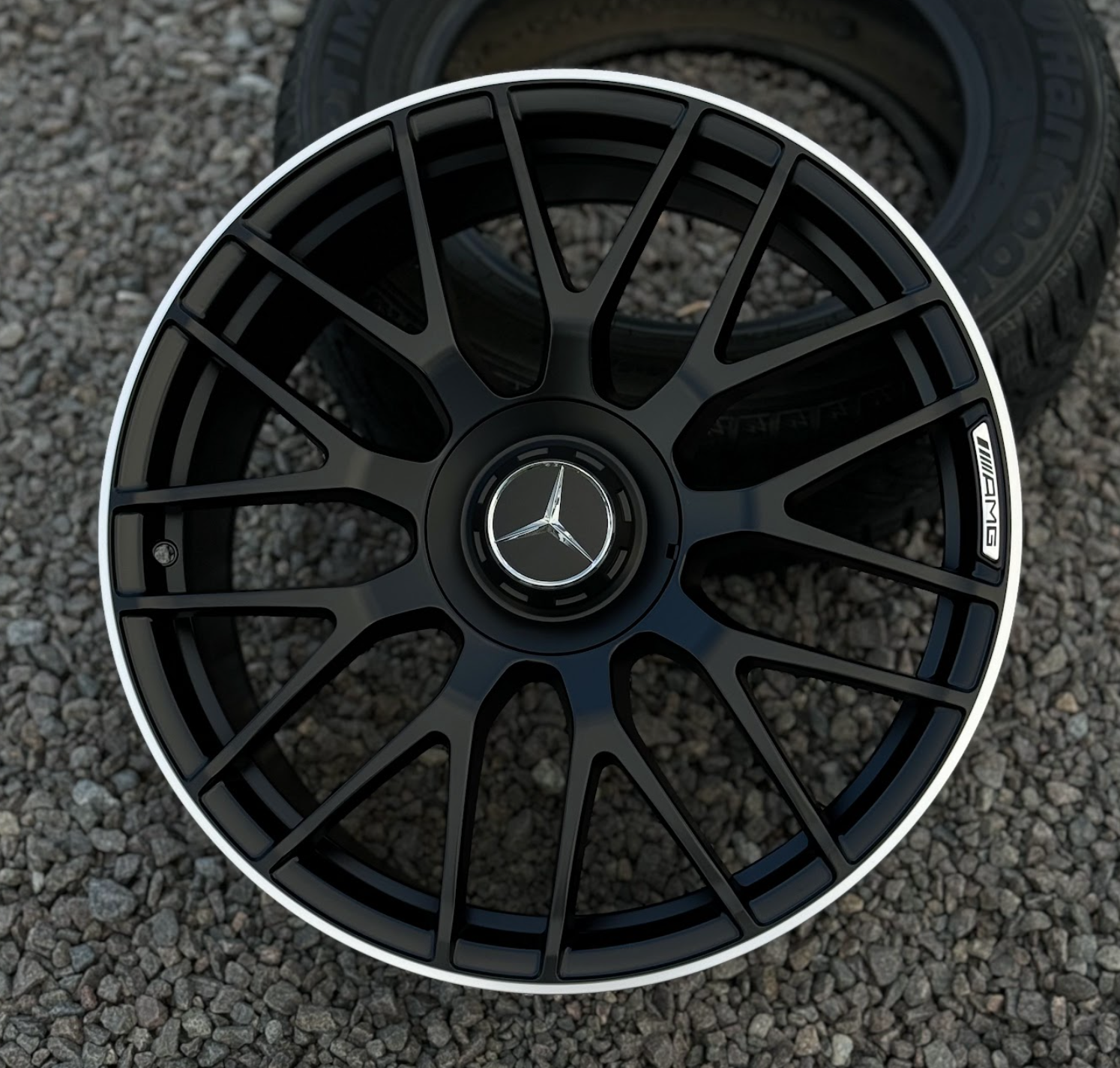 racingline_rfx-146_blackmachinedface Close-up of a black multi-spoke Mercedes-AMG alloy wheel with a silver rim and central Mercedes emblem, resting on gravel.