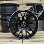 Close-up of a black Mercedes‑AMG alloy wheel with a silver lip, against a wooden pallet backdrop by stacked rims.