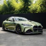 Front three-quarter view of a lime-green BMW sports car with black alloy wheels parked in front of a dark wooden fence and dense green trees behind it.