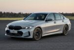 Front three-quarter view of a silver BMW sedan on an open road with a grassy field in the background, during sunset.