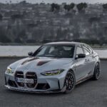 Silver BMW sports sedan with black and red racing stripes, parked on a paved road by a body of water.