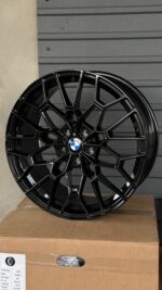 Glossy black BMW alloy wheel with an intricate spoke design and center BMW emblem, resting on a cardboard box.