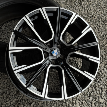 Close-up of a black and silver BMW alloy wheel with bold spokes on a gravel surface, center cap with BMW logo visible.