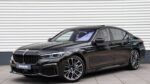 Front three-quarter view of a black BMW sedan parked on a light-gray tiled surface in front of a corrugated metal wall.