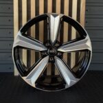 Close-up of a glossy Audi alloy wheel with black and silver spokes, center hub, and logo visible, set against a wooden backdrop and dark floor.