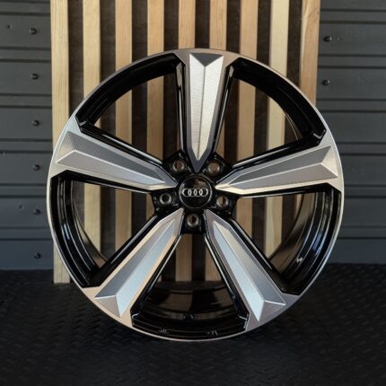 Close-up of a glossy Audi alloy wheel with black and silver spokes, center hub, and logo visible, set against a wooden backdrop and dark floor.