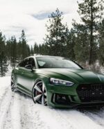 Green Audi sedan parked on a snow-covered forest road with evergreen trees in the background.