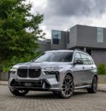 Three-quarter front view of a silver BMW SUV parked on a brick driveway in front of a modern gray building with trees nearby.