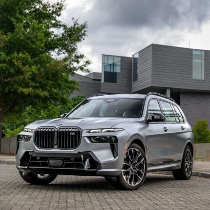 Three-quarter front view of a silver BMW SUV parked on a brick driveway in front of a modern gray building with trees nearby.