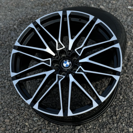 BMW alloy wheel with black and silver geometric spokes on a gravel surface, centered BMW logo.