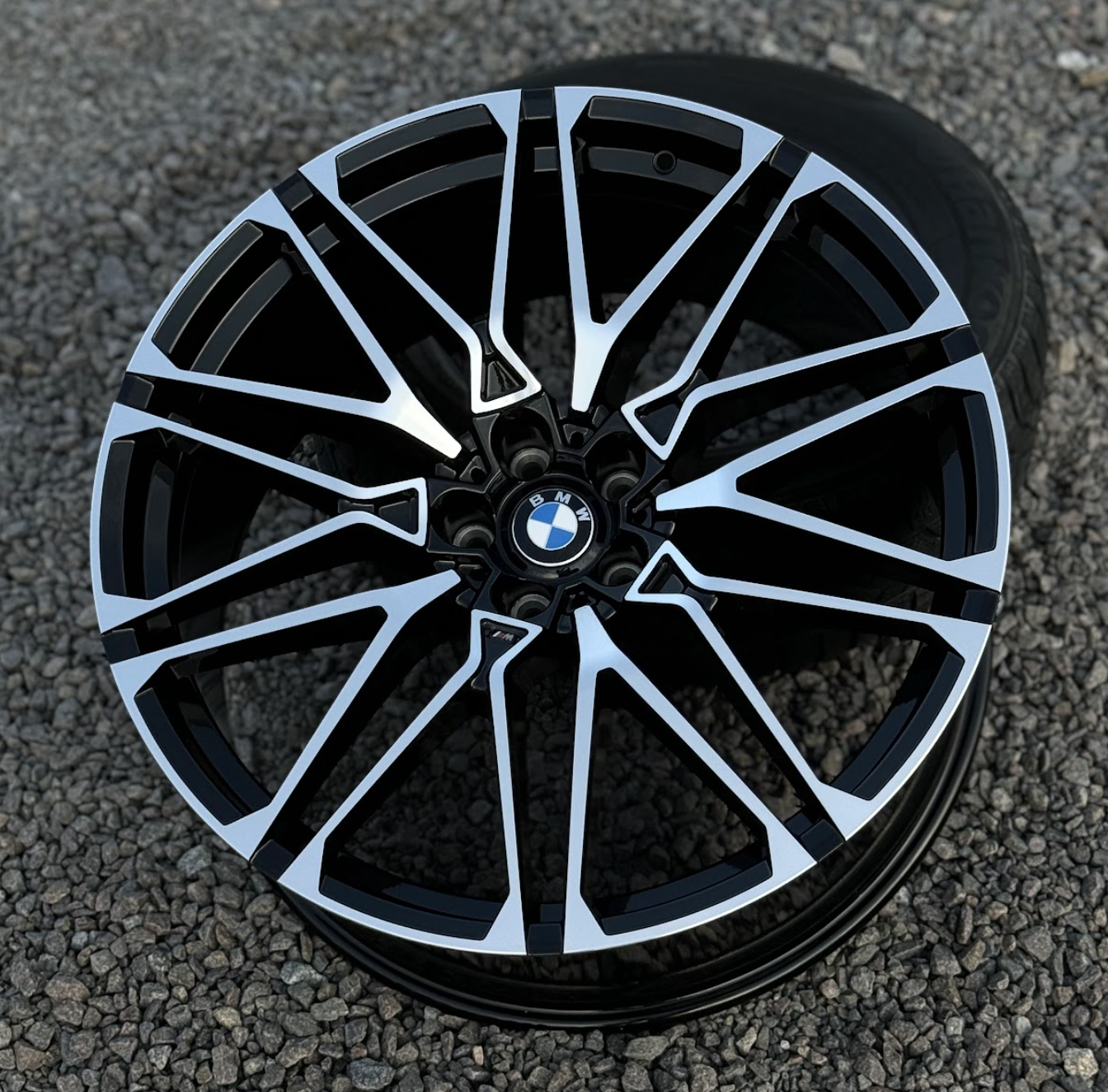 racingline_vfx-495_blackmachinedface BMW alloy wheel with black and silver geometric spokes on a gravel surface, centered BMW logo.